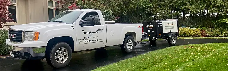 White pickup truck with company branding "Ronin Omar Inc" towing a black and white industrial trailer on a driveway, surrounded by green lawn and trees.