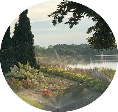 A sprinkler waters a lush lawn in front of a serene lake, creating a picturesque outdoor scene.