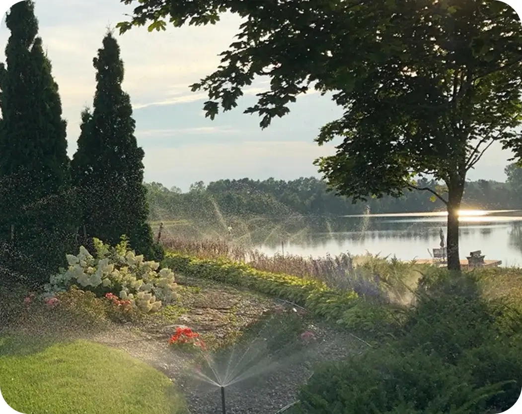 A sprinkler waters a lush lawn in front of a serene lake, creating a picturesque outdoor scene.